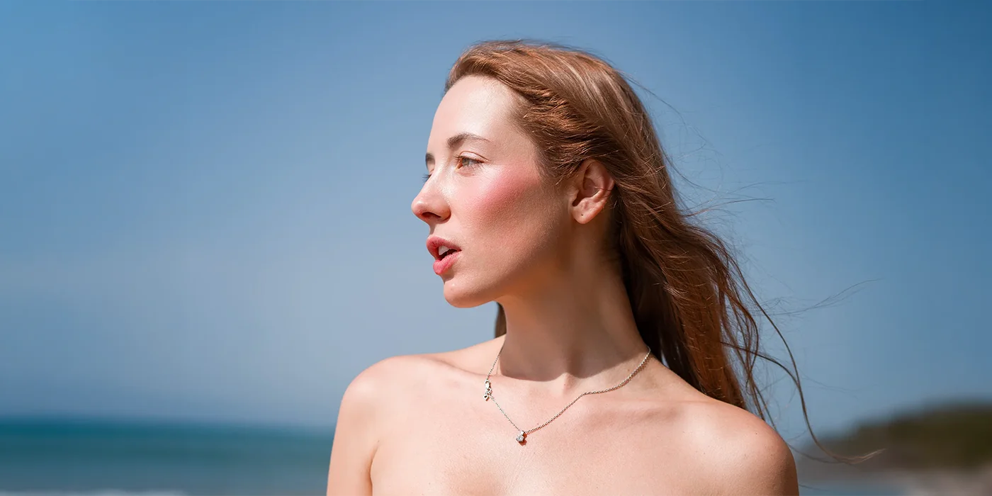 Close-up portrait of a young woman with auburn hair and a delicate silver necklace, photographed outdoors in natural sunlight against a blurred ocean and blue sky background